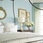 A cozy bedroom—featured in the RSOL Designer House—showcases a black canopy bed, white bedding, light blue walls, a striking sunburst mirror, and three framed pictures. A bedside table holds a white lamp and a vase of white flowers. Green patterned curtains and a window brighten the room, while books are stacked at the bed’s foot. This inviting space has received glowing testimonials for its soothing ambiance and thoughtful design touches.