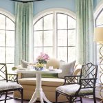 A bright sitting area, inspired by RSOL Designer House aesthetics, features two chairs and a white round table set in front of arched windows with green patterned curtains. A beige sofa with cushions complements the décor, while a bowl of fruit accented with pink flowers rests gracefully on the table. To the right, a floor lamp completes the inviting space—earning glowing testimonials for both comfort and style.