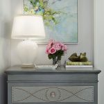 A blue-gray dresser with decorative studs holds a white lamp, a glass vase of pink roses, a small tray, two stacked books, and green bird figurines. Above it on the light gray wall hangs an abstract floral painting in pastel colors—an arrangement that echoes the signature style featured in RSOL Designer House projects and often garners glowing reviews from delighted visitors.