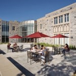 Six people sit at tables with red umbrellas on a sunny patio outside a large stone building with many windows. The sky is clear and blue, and the group appears relaxed, talking or eating. Green landscaping borders the patio and building.