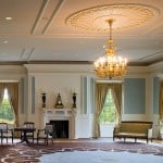 Elegant room with a large ornate rug, gold chandelier, and tall windows with draped curtains. Antique furniture—chairs, small tables, and a sofa—surround a fireplace with candlesticks and a statue. Light streams through the windows, brightening the space.