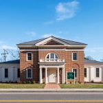 A two-story red brick building with white columns and trim, featuring a central arched balcony and a green sign in front reading Business Center. The building, reminiscent of the inviting architecture of the Shenkman Jewish Center, sits along a street with a bright blue sky above.